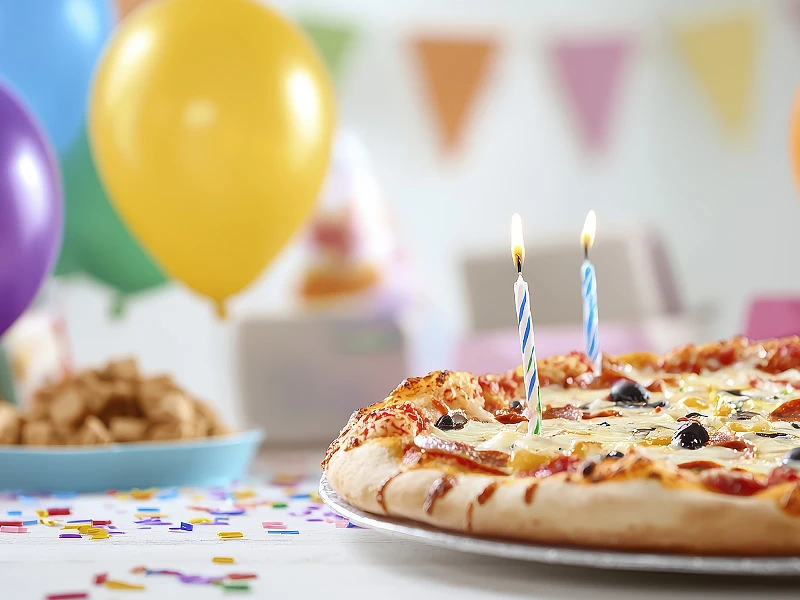 Birthday celebration scene with a pizza decorated with two lit candles, surrounded by balloons and party decorations.